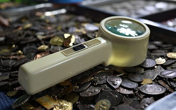 DFC 4172 A handheld magnifying glass resting on a scattered pile of coins and medallions highlighting intricat