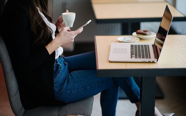Woman sitting in cafe enjoying coffee and using smartphone while laptop rests on table